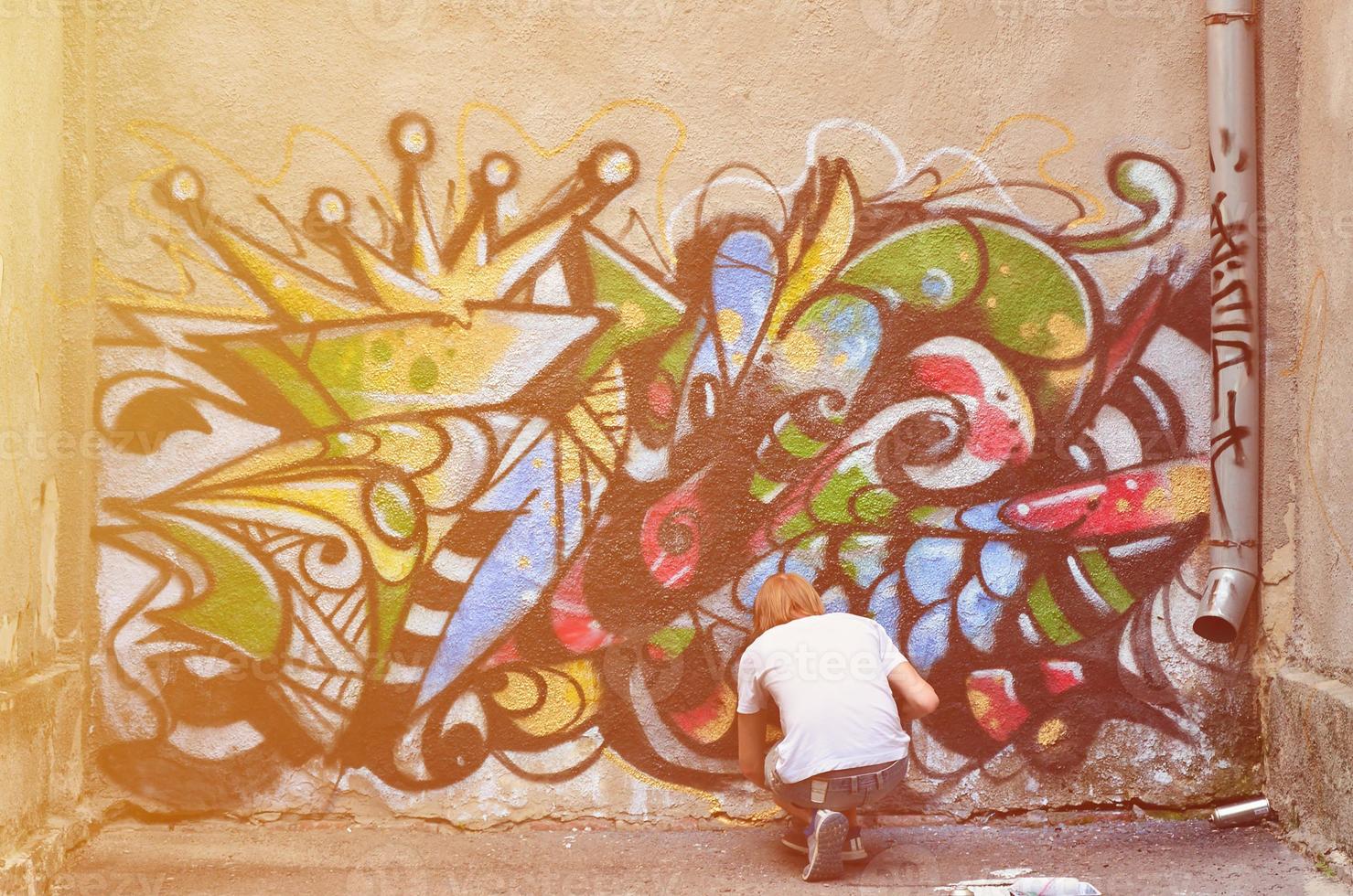 foto en el proceso de dibujar un patrón de graffiti en un antiguo muro de hormigón. un joven rubio de pelo largo dibuja un dibujo abstracto de diferentes colores. concepto de arte callejero y vandalismo