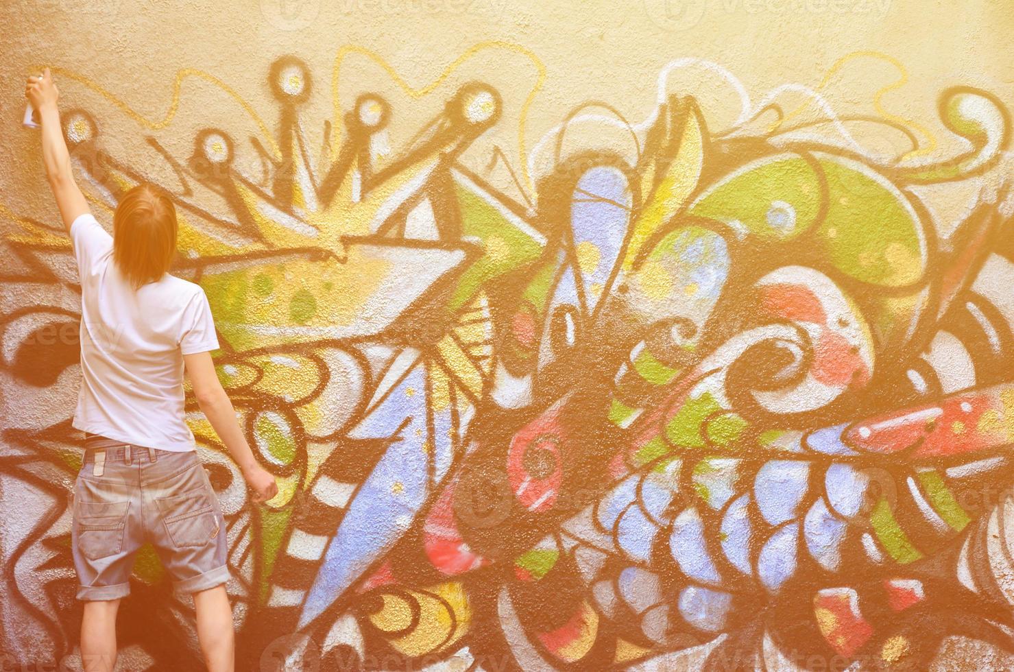 Photo of a young guy in denim shorts and a white shirt. The guy draws on the graffiti wall a drawing with aerosol paints of various colors. The concept of hooliganism and damage to property