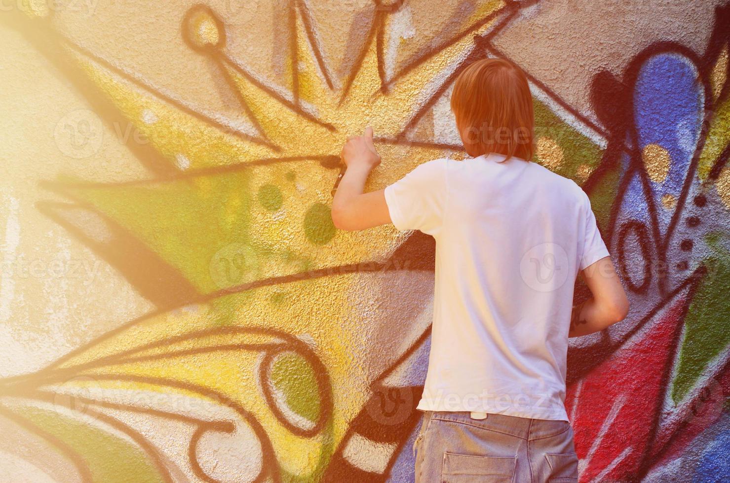 Photo of a young guy in denim shorts and a white shirt. The guy draws on the graffiti wall a drawing with aerosol paints of various colors. The concept of hooliganism and damage to property