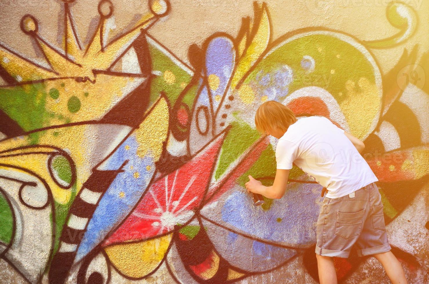 foto de un joven con pantalones cortos de mezclilla y una camisa blanca. el chico dibuja en la pared de graffiti un dibujo con pinturas en aerosol de varios colores. el concepto de vandalismo y daños a la propiedad