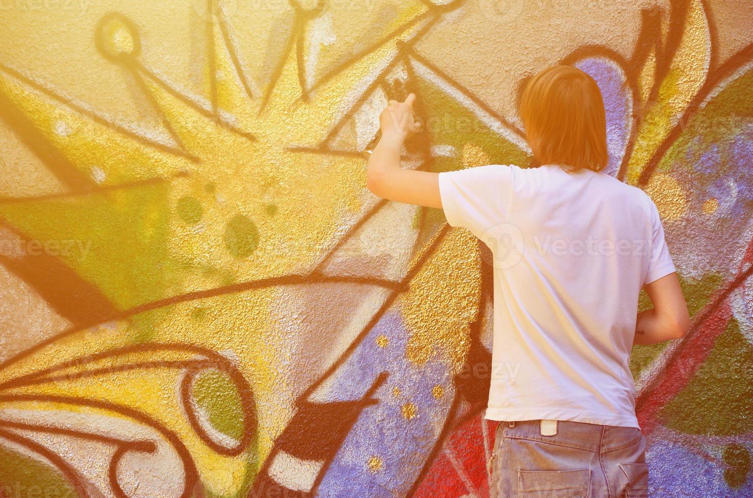 Photo in the process of drawing a graffiti pattern on an old concrete wall. Young long-haired blond guy draws an abstract drawing of different colors. Street art and vandalism concept
