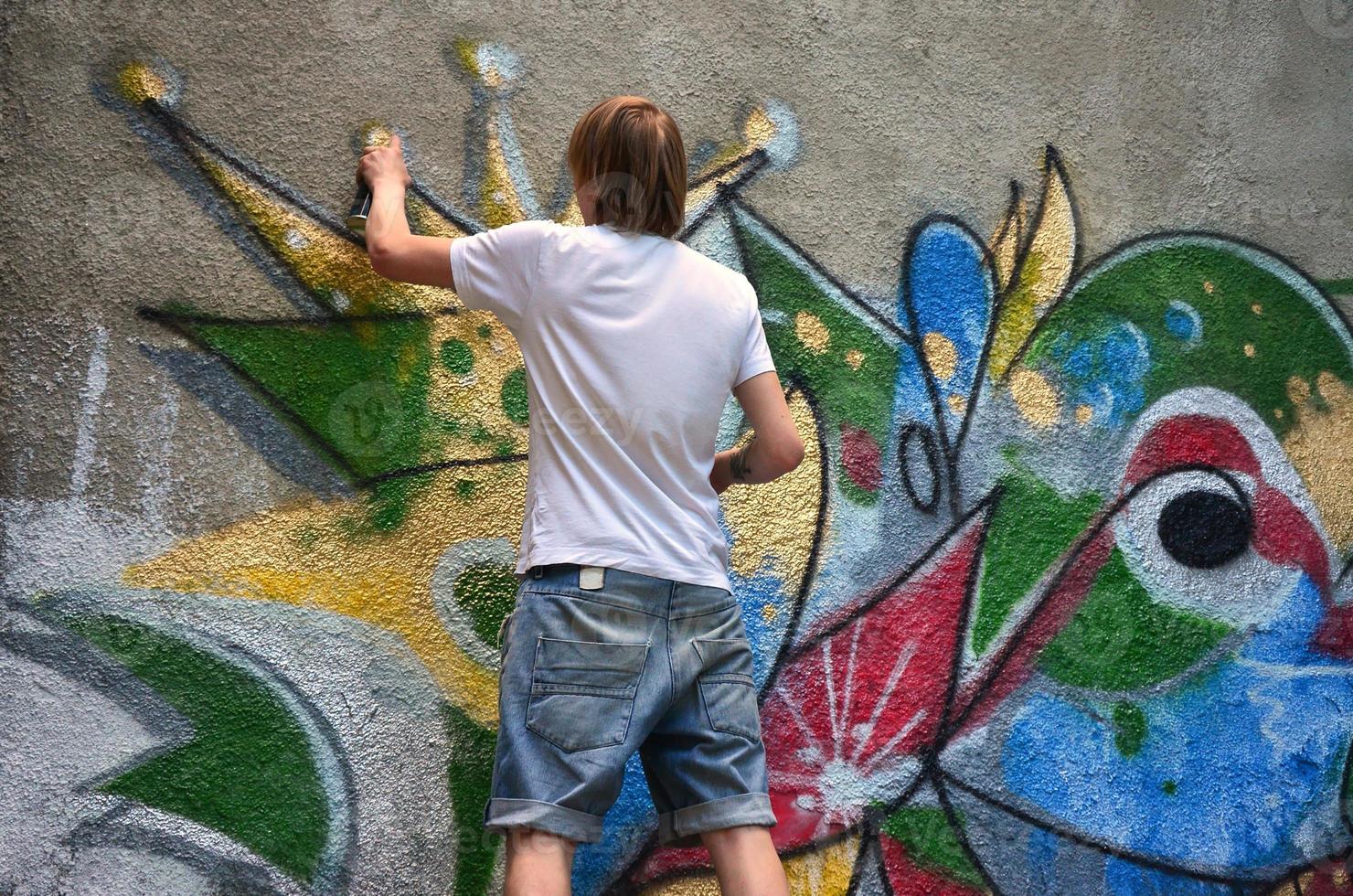 Photo of a young guy in denim shorts and a white shirt. The guy draws on the graffiti wall a drawing with aerosol paints of various colors. The concept of hooliganism and damage to property