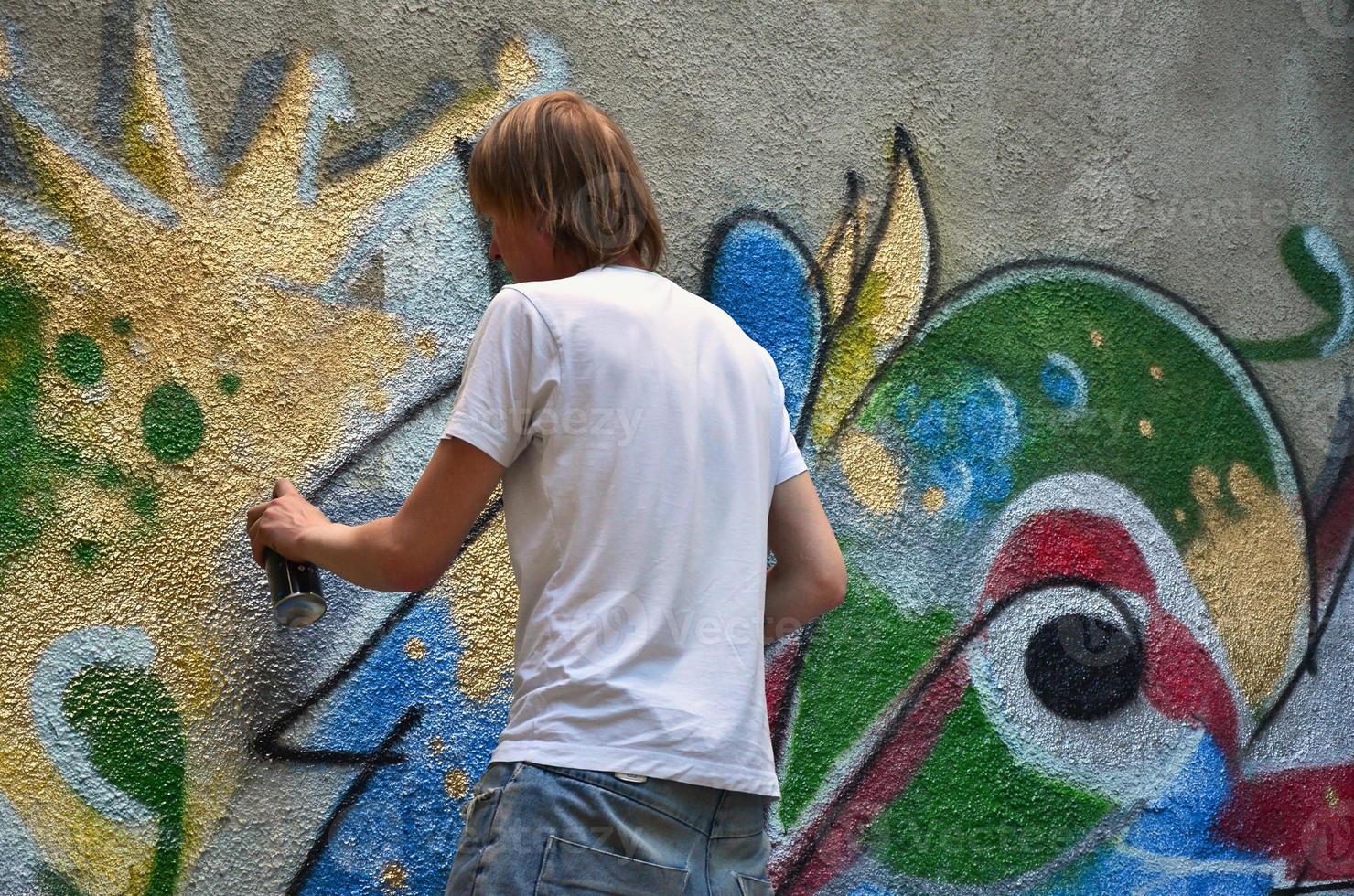 Photo in the process of drawing a graffiti pattern on an old concrete wall. Young long-haired blond guy draws an abstract drawing of different colors. Street art and vandalism concept