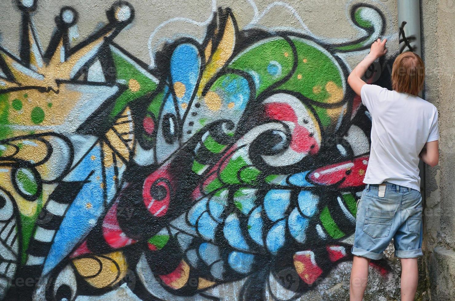 Photo in the process of drawing a graffiti pattern on an old concrete wall. Young long-haired blond guy draws an abstract drawing of different colors. Street art and vandalism concept