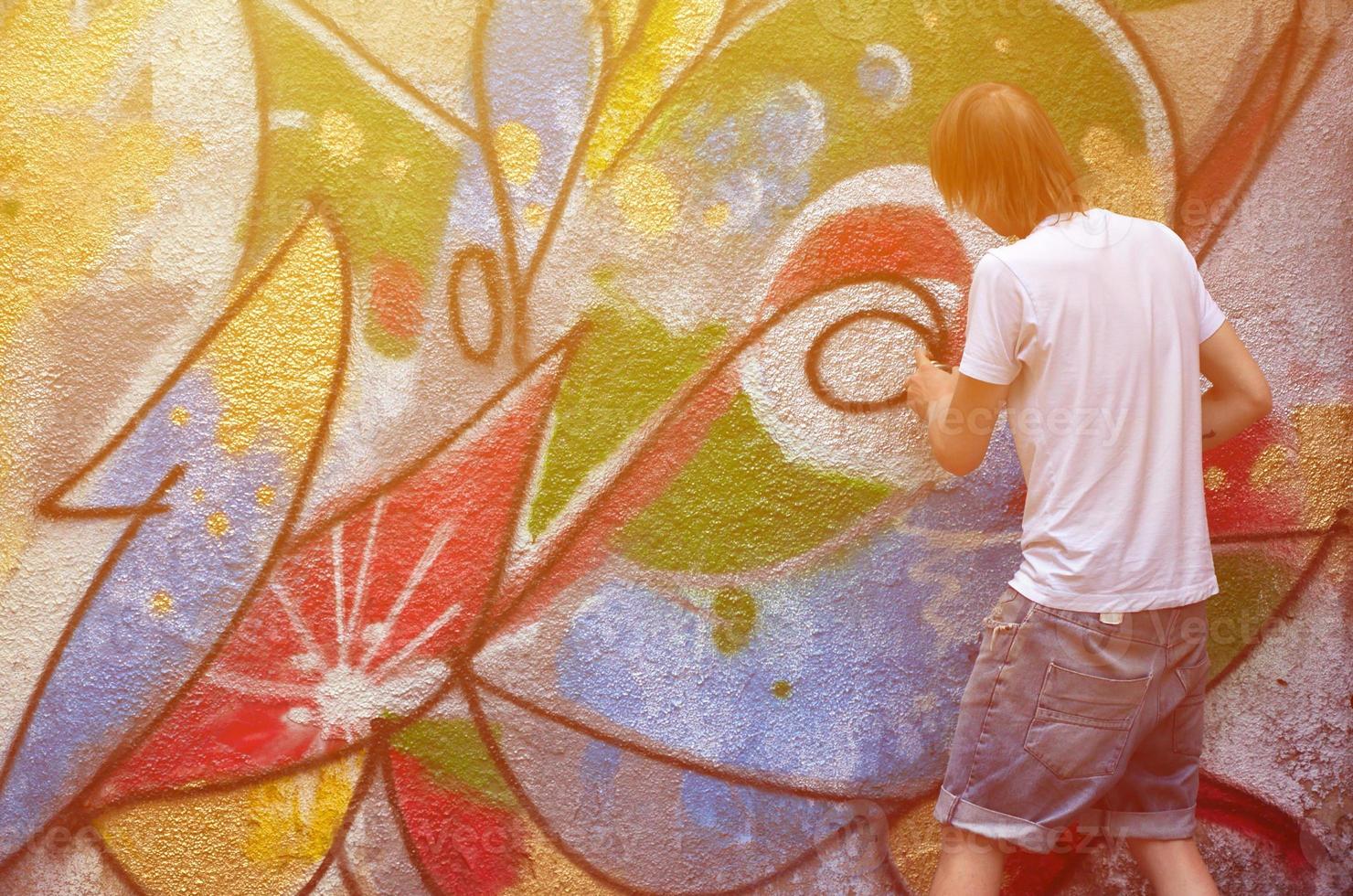 foto en el proceso de dibujar un patrón de graffiti en un antiguo muro de hormigón. un joven rubio de pelo largo dibuja un dibujo abstracto de diferentes colores. concepto de arte callejero y vandalismo