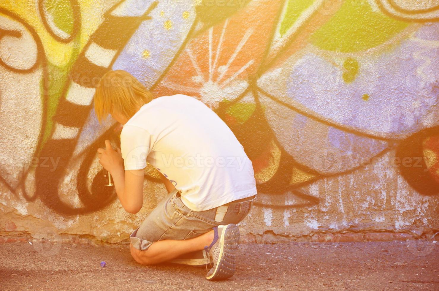 Photo in the process of drawing a graffiti pattern on an old concrete wall. Young long-haired blond guy draws an abstract drawing of different colors. Street art and vandalism concept
