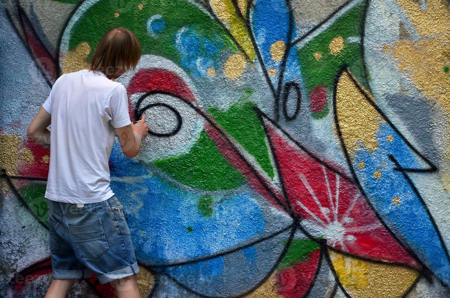 Photo in the process of drawing a graffiti pattern on an old concrete wall. Young long-haired blond guy draws an abstract drawing of different colors. Street art and vandalism concept