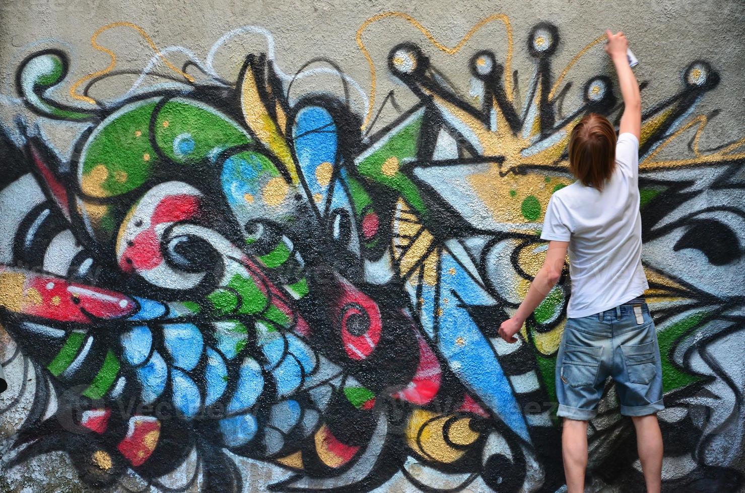 Photo in the process of drawing a graffiti pattern on an old concrete wall. Young long-haired blond guy draws an abstract drawing of different colors. Street art and vandalism concept
