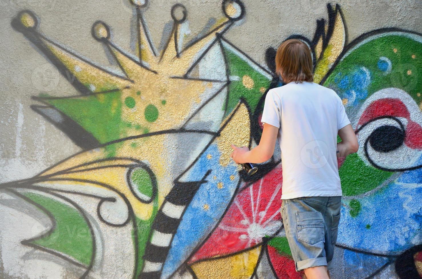 Photo in the process of drawing a graffiti pattern on an old concrete wall. Young long-haired blond guy draws an abstract drawing of different colors. Street art and vandalism concept