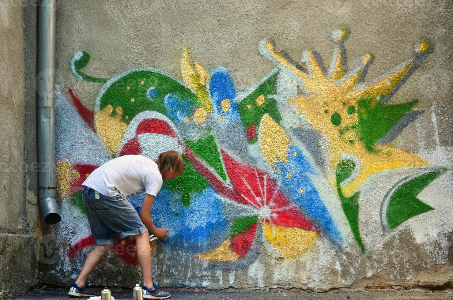 Photo of a young guy in denim shorts and a white shirt. The guy draws on the graffiti wall a drawing with aerosol paints of various colors. The concept of hooliganism and damage to property