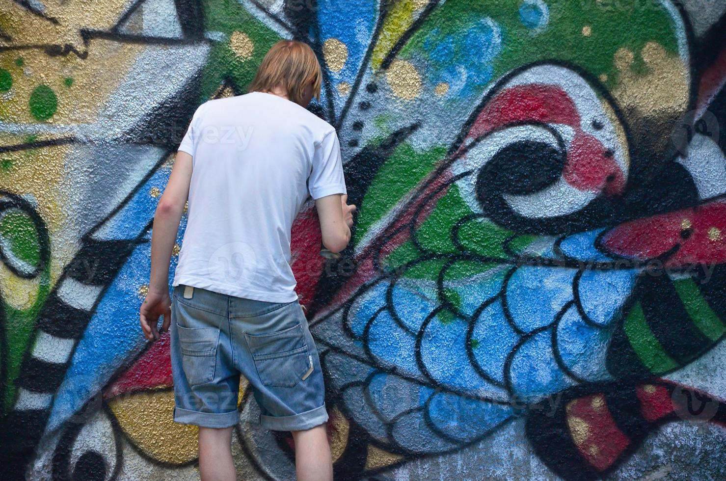 Photo in the process of drawing a graffiti pattern on an old concrete wall. Young long-haired blond guy draws an abstract drawing of different colors. Street art and vandalism concept