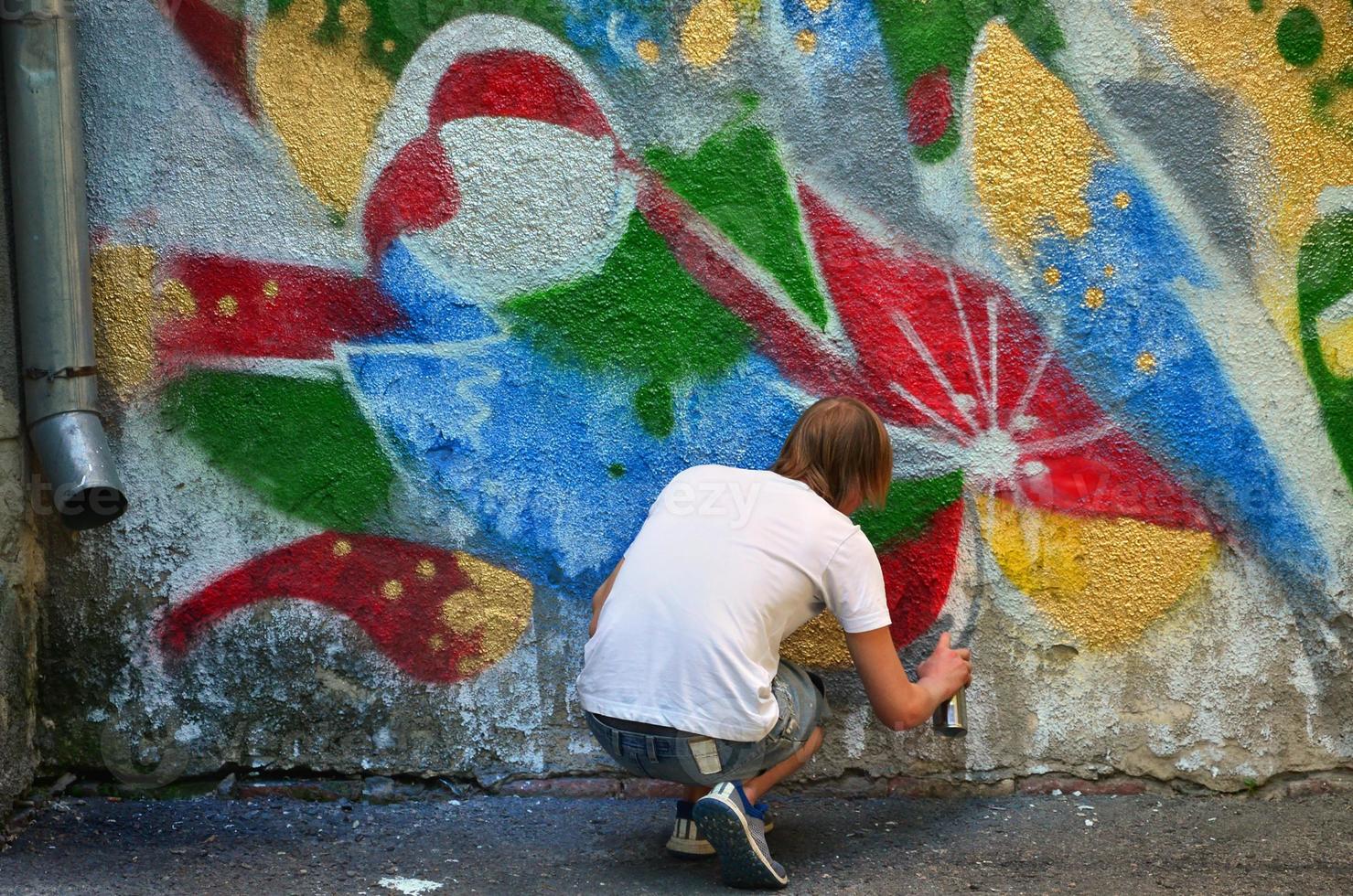 Photo of a young guy in denim shorts and a white shirt. The guy draws on the graffiti wall a drawing with aerosol paints of various colors. The concept of hooliganism and damage to property