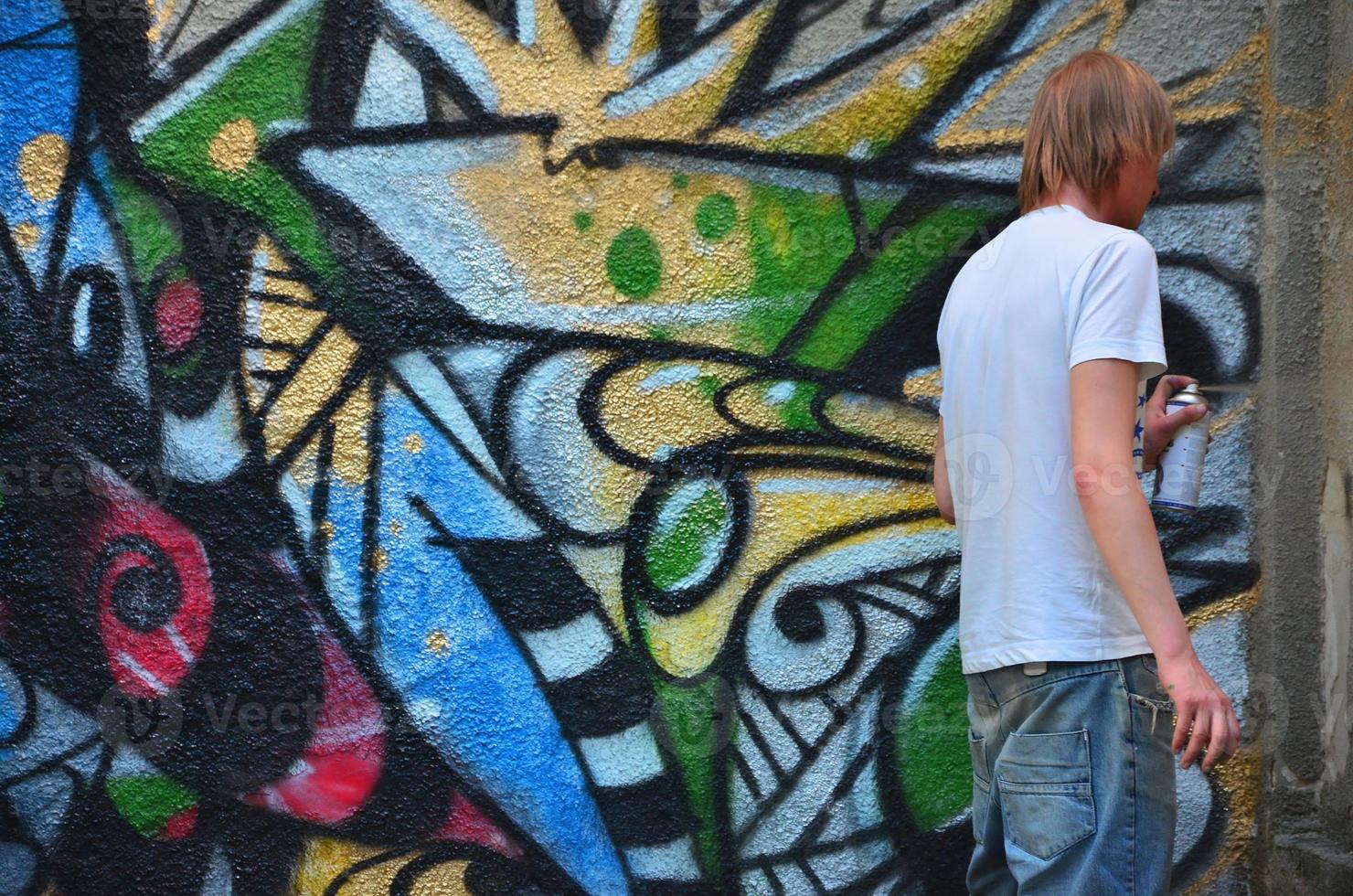 Photo in the process of drawing a graffiti pattern on an old concrete wall. Young long-haired blond guy draws an abstract drawing of different colors. Street art and vandalism concept