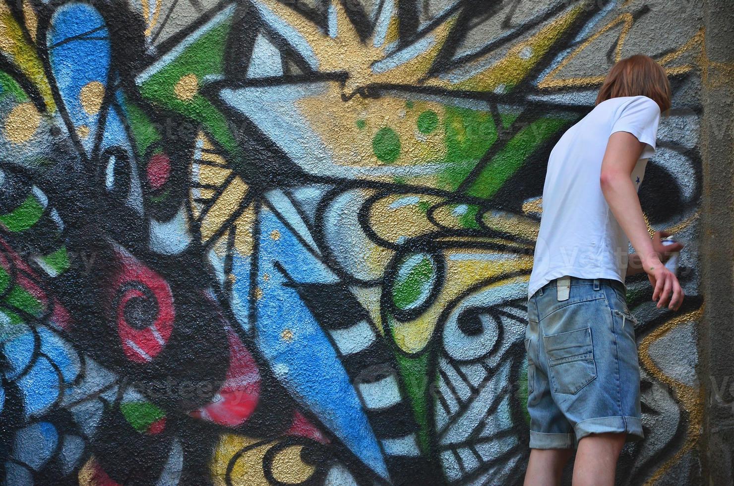 Photo in the process of drawing a graffiti pattern on an old concrete wall. Young long-haired blond guy draws an abstract drawing of different colors. Street art and vandalism concept
