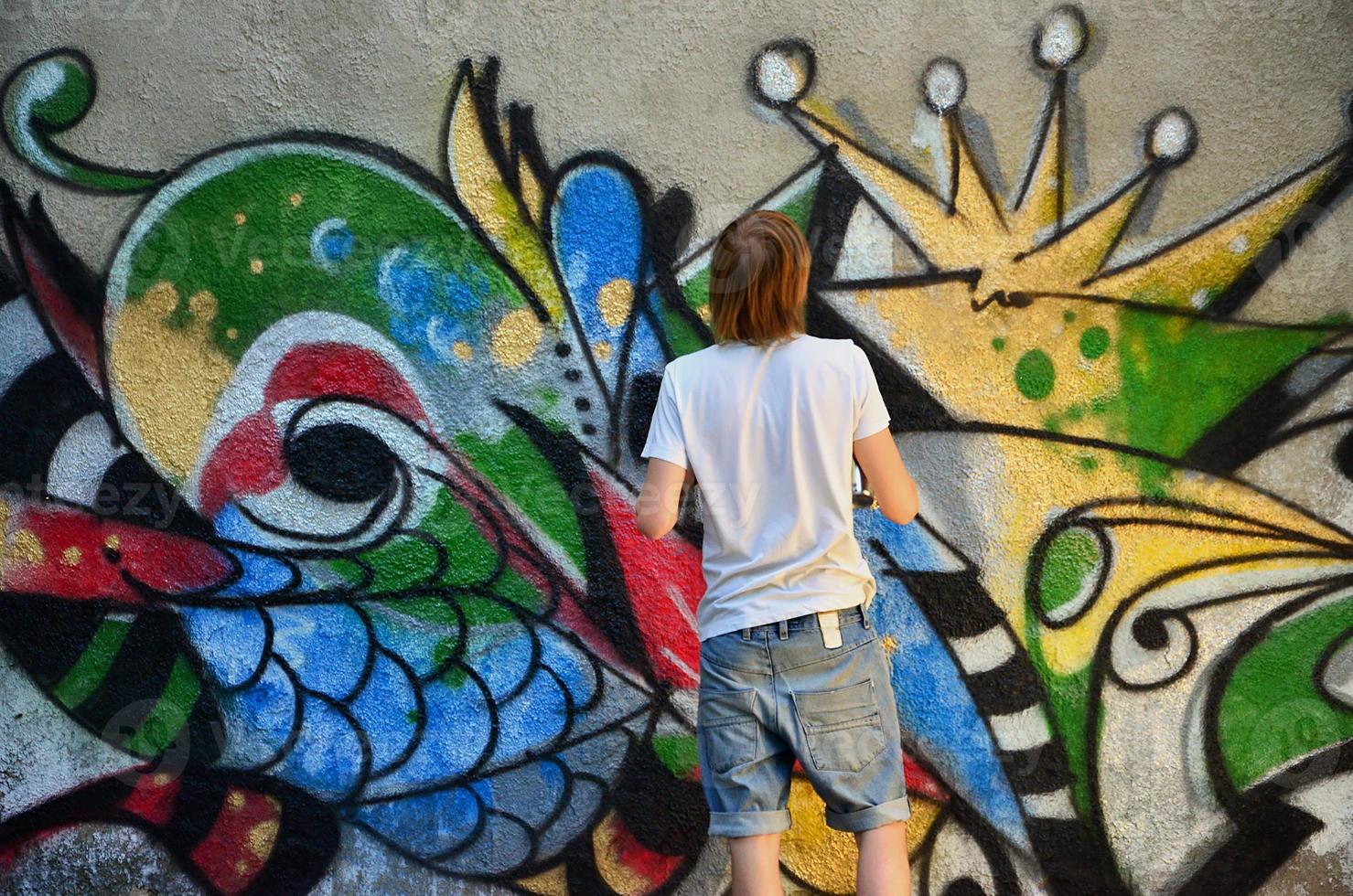 Photo in the process of drawing a graffiti pattern on an old concrete wall. Young long-haired blond guy draws an abstract drawing of different colors. Street art and vandalism concept