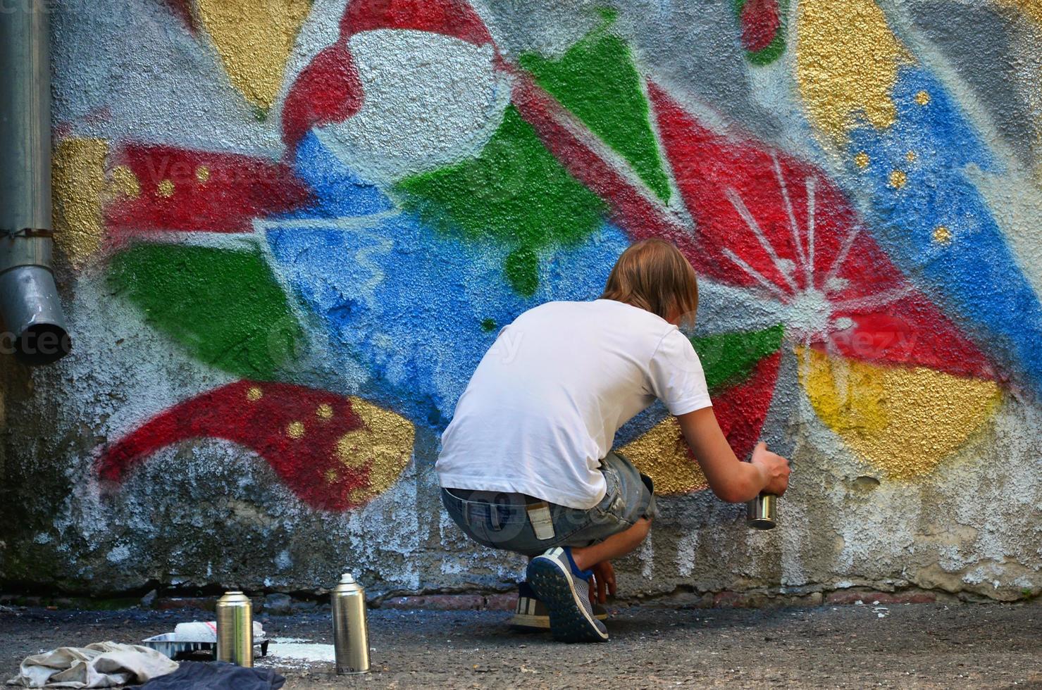 Photo in the process of drawing a graffiti pattern on an old concrete wall. Young long-haired blond guy draws an abstract drawing of different colors. Street art and vandalism concept