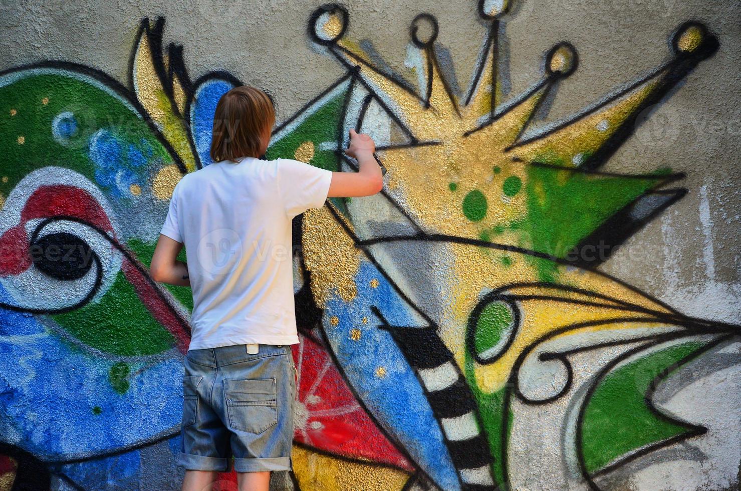 Photo of a young guy in denim shorts and a white shirt. The guy draws on the graffiti wall a drawing with aerosol paints of various colors. The concept of hooliganism and damage to property