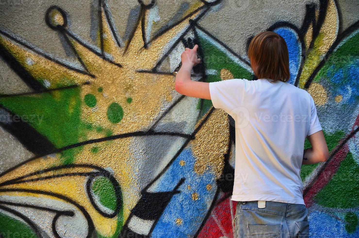 foto en el proceso de dibujar un patrón de graffiti en un antiguo muro de hormigón. un joven rubio de pelo largo dibuja un dibujo abstracto de diferentes colores. concepto de arte callejero y vandalismo