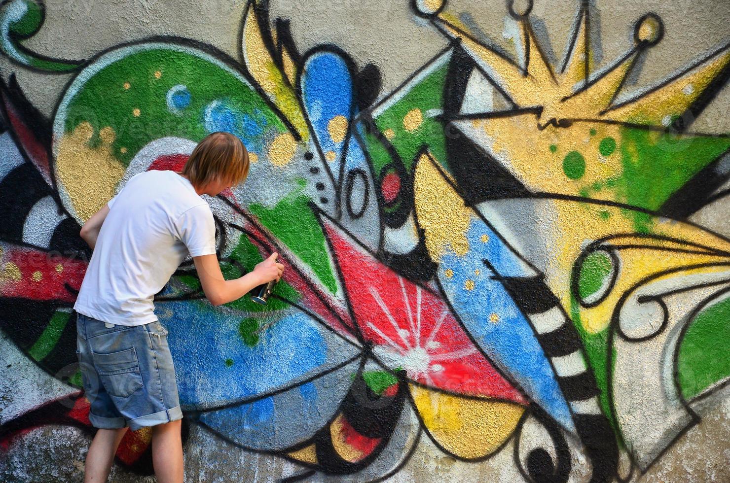 Photo in the process of drawing a graffiti pattern on an old concrete wall. Young long-haired blond guy draws an abstract drawing of different colors. Street art and vandalism concept
