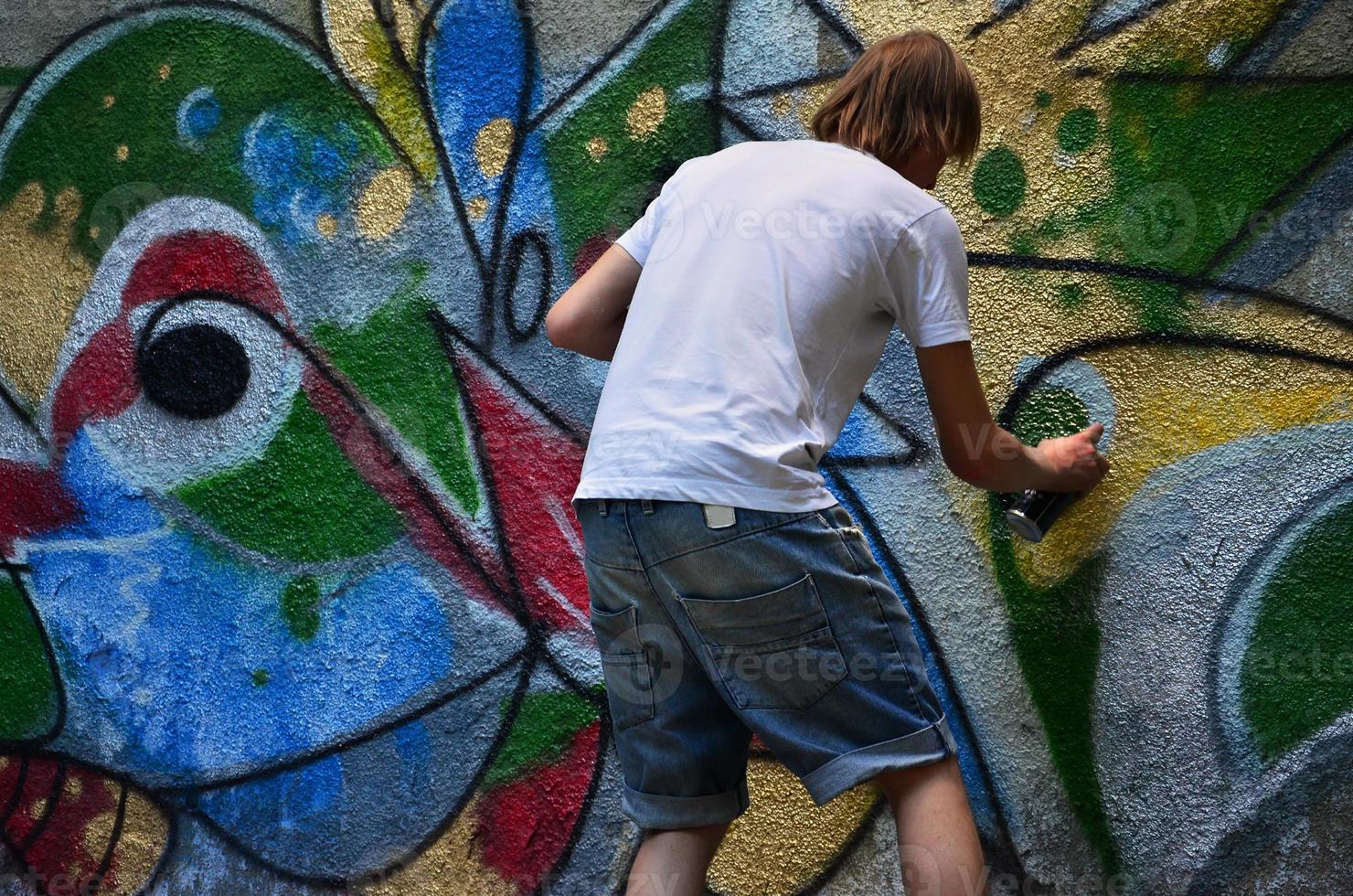 foto en el proceso de dibujar un patrón de graffiti en un antiguo muro de hormigón. un joven rubio de pelo largo dibuja un dibujo abstracto de diferentes colores. concepto de arte callejero y vandalismo