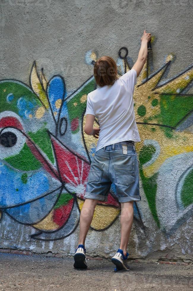 Photo in the process of drawing a graffiti pattern on an old concrete wall. Young long-haired blond guy draws an abstract drawing of different colors. Street art and vandalism concept