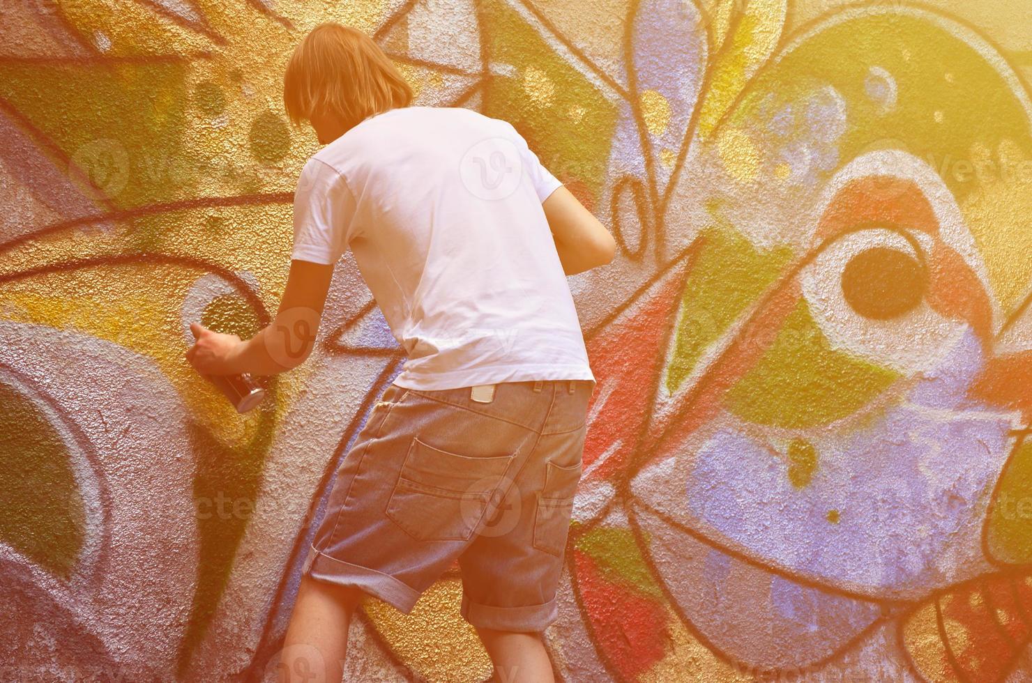 Photo in the process of drawing a graffiti pattern on an old concrete wall. Young long-haired blond guy draws an abstract drawing of different colors. Street art and vandalism concept