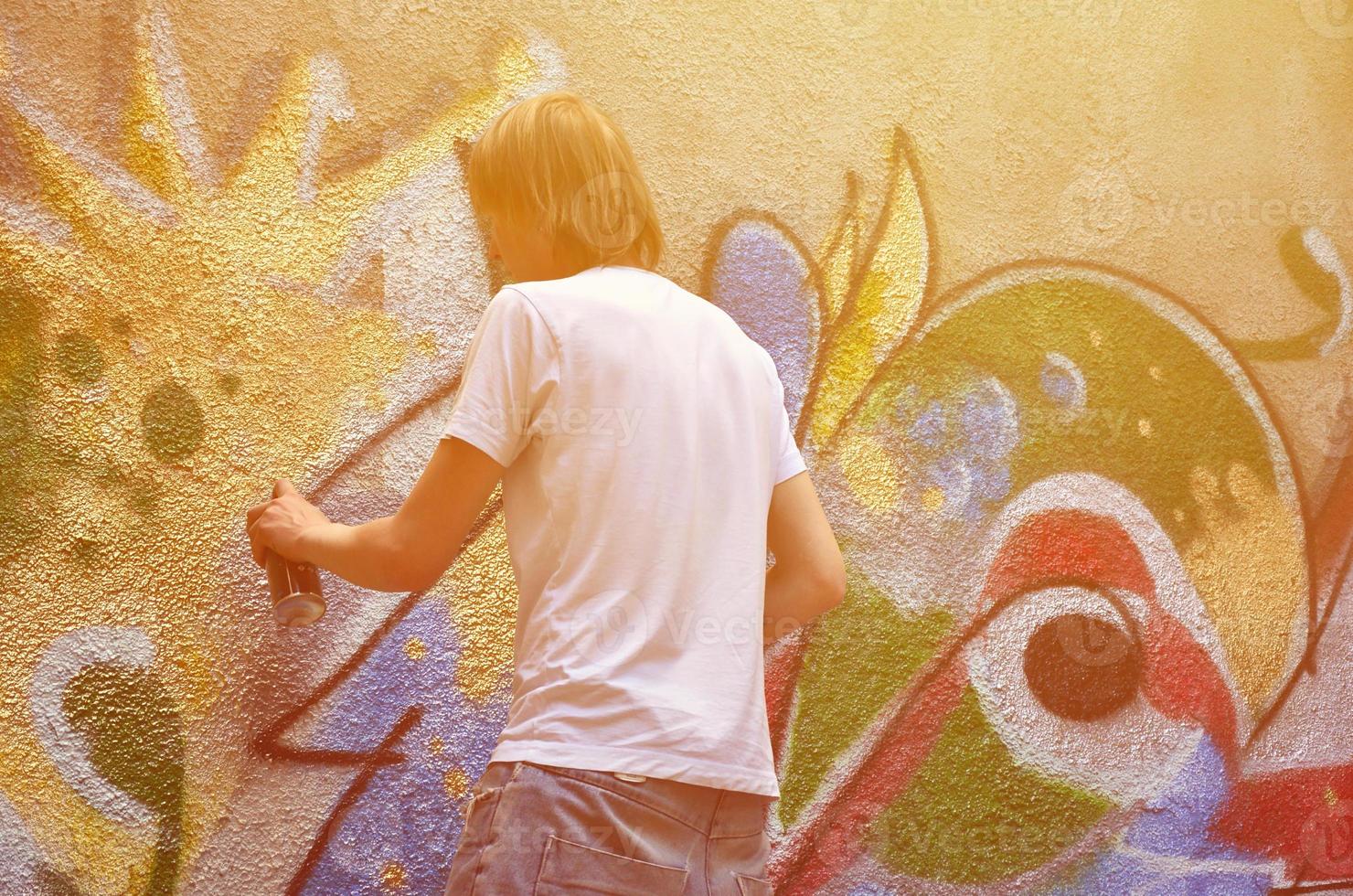 Photo in the process of drawing a graffiti pattern on an old concrete wall. Young long-haired blond guy draws an abstract drawing of different colors. Street art and vandalism concept