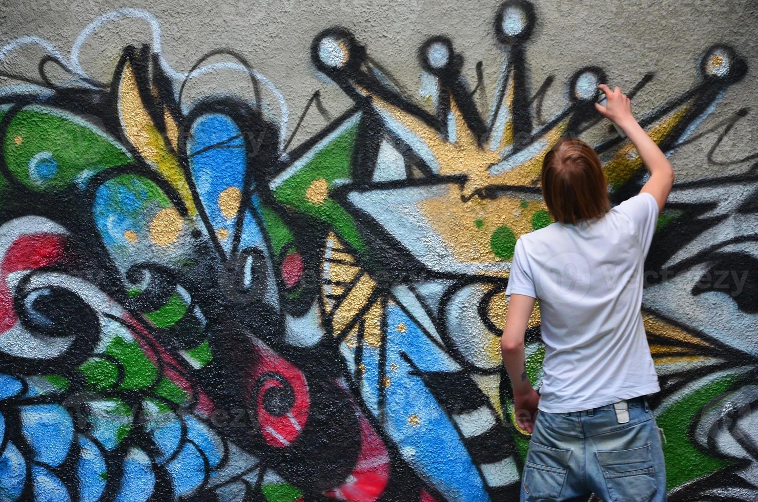 Photo in the process of drawing a graffiti pattern on an old concrete wall. Young long-haired blond guy draws an abstract drawing of different colors. Street art and vandalism concept