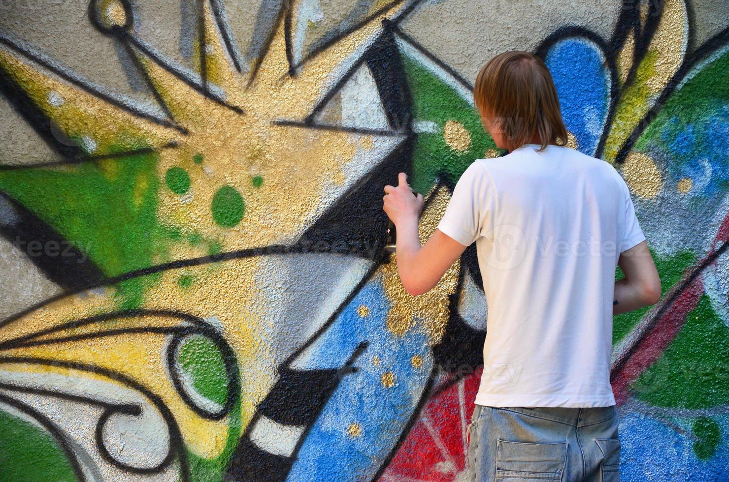 Photo in the process of drawing a graffiti pattern on an old concrete wall. Young long-haired blond guy draws an abstract drawing of different colors. Street art and vandalism concept
