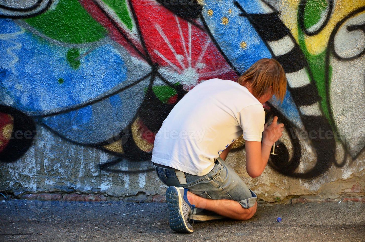 Photo in the process of drawing a graffiti pattern on an old concrete wall. Young long-haired blond guy draws an abstract drawing of different colors. Street art and vandalism concept