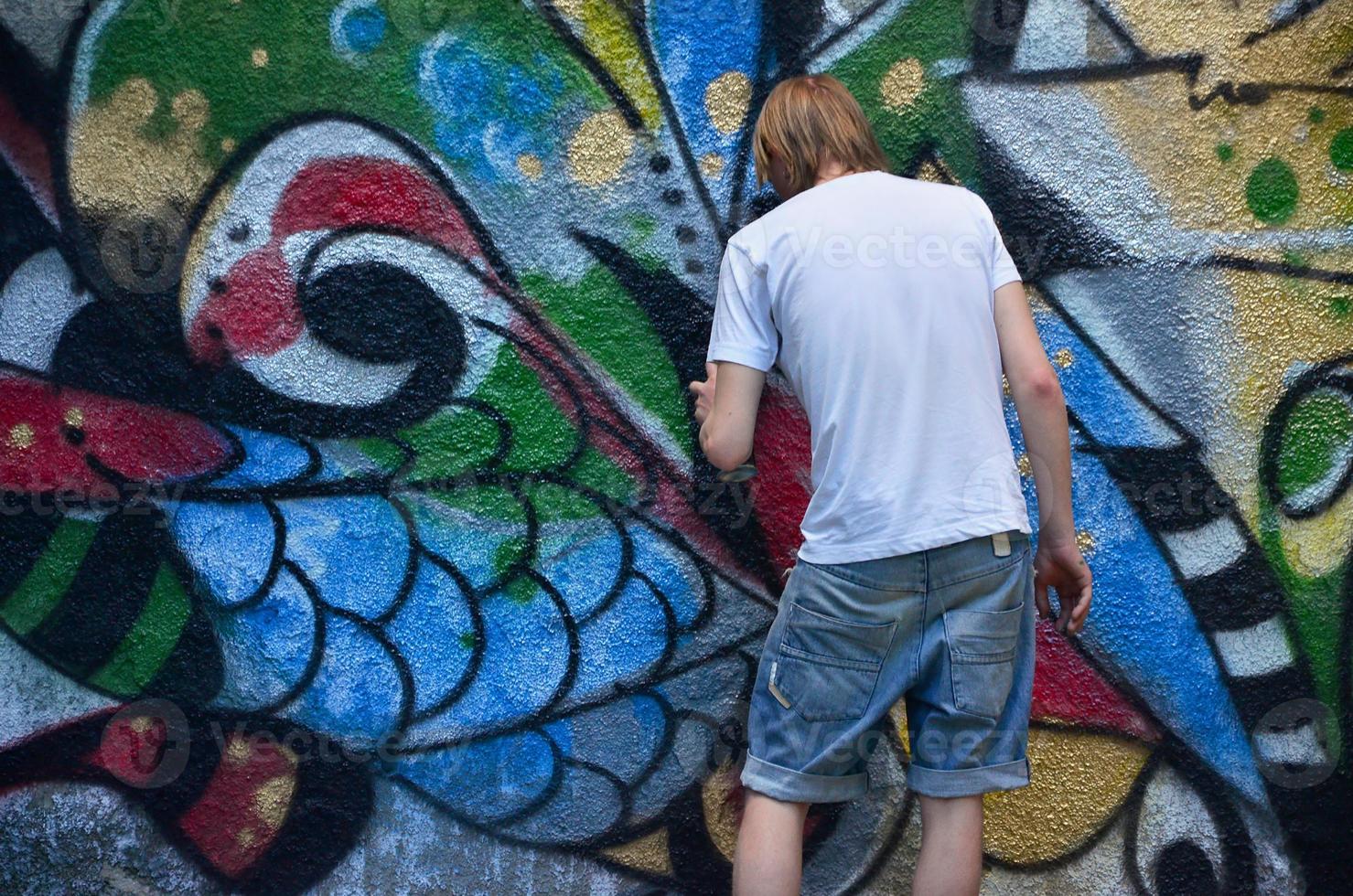 Photo in the process of drawing a graffiti pattern on an old concrete wall. Young long-haired blond guy draws an abstract drawing of different colors. Street art and vandalism concept