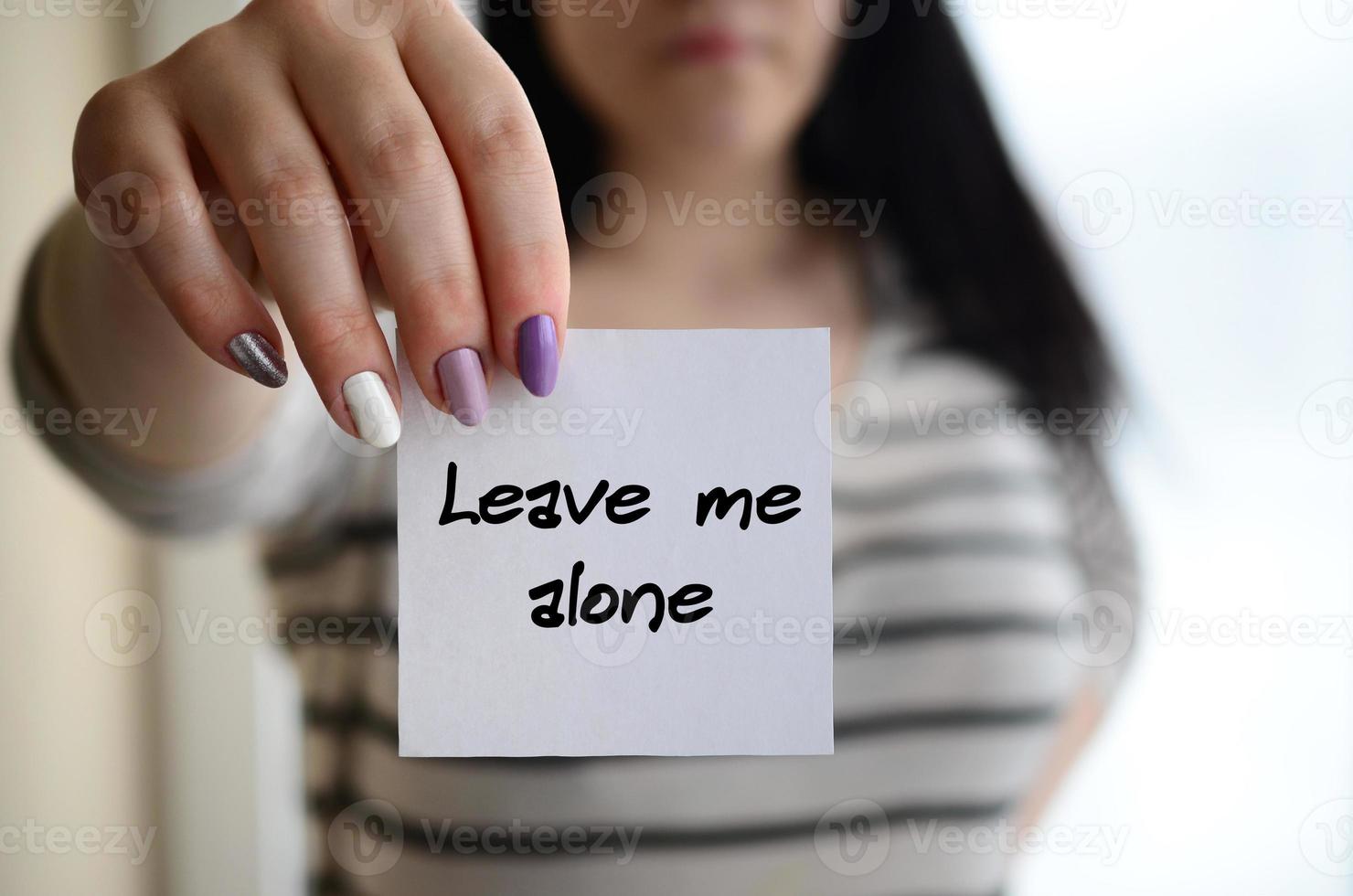 Young sad girl shows a white sticker. Caucasian brunette holding a sheet of paper with message. Leave me alone photo