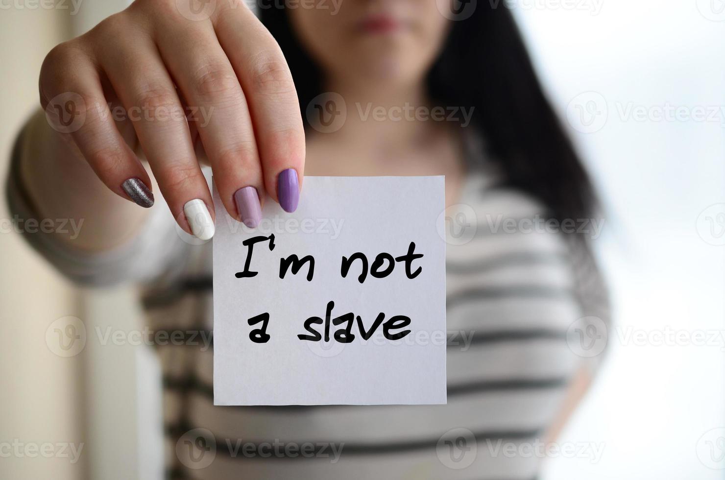 Young sad girl shows a white sticker. Caucasian brunette holding a sheet of paper with message. Im not a slave photo