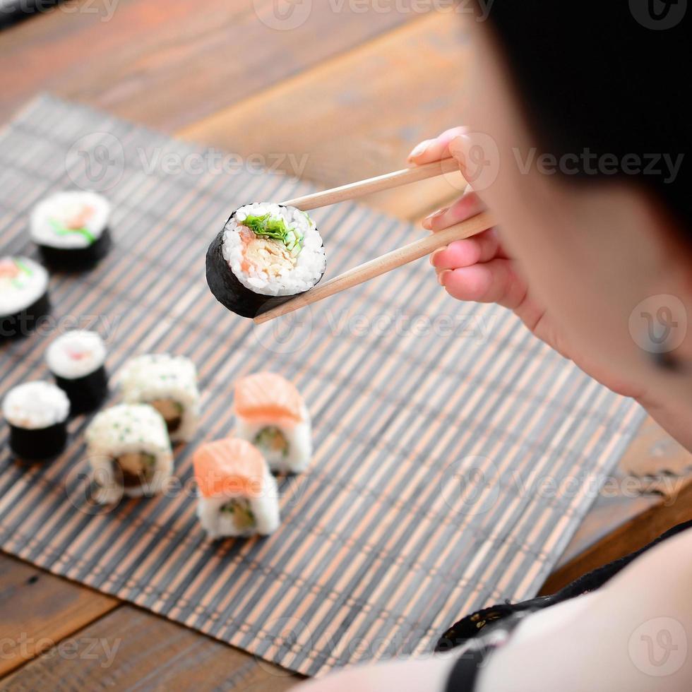 una chica morena con palillos sostiene un rollo de sushi sobre un fondo de alfombra de paja de bambú. comida asiática tradicional foto