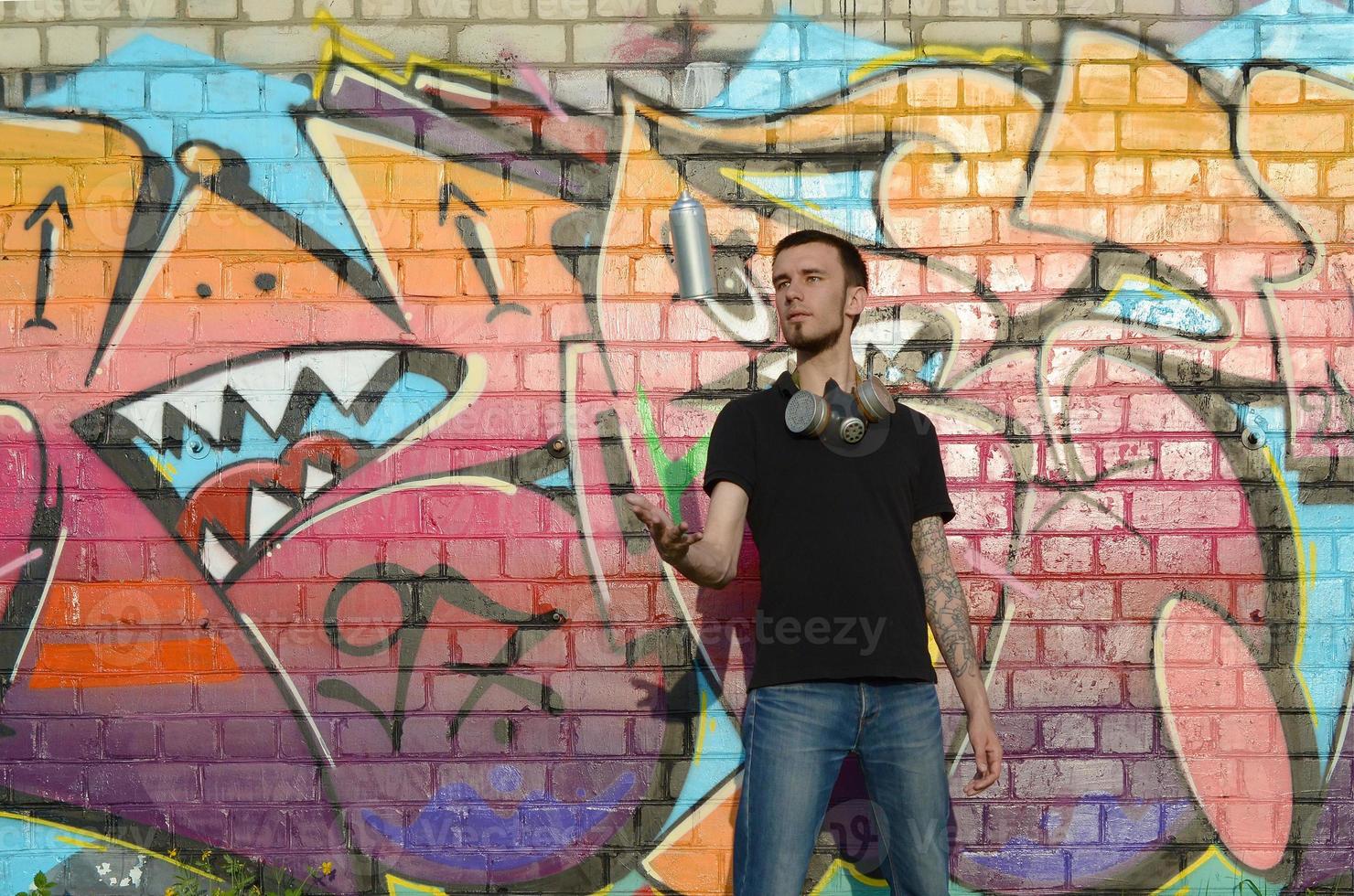 Young graffiti artist with gas mask on his neck throw his spray can against colorful pink graffiti on brick wall. Street art and contemporary painting process photo