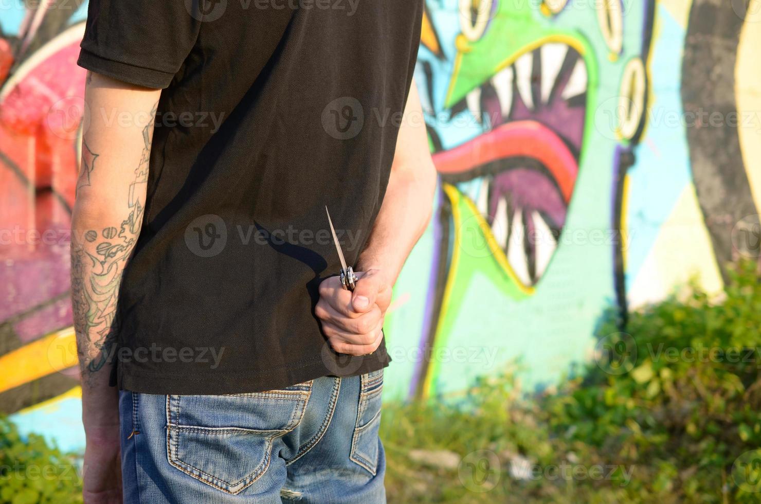 vista trasera de un joven caucásico con un cuchillo en la mano contra la pared de ladrillo del ghetto con pinturas de graffiti. concepto de fuerzas criminales y cargo de agresión foto