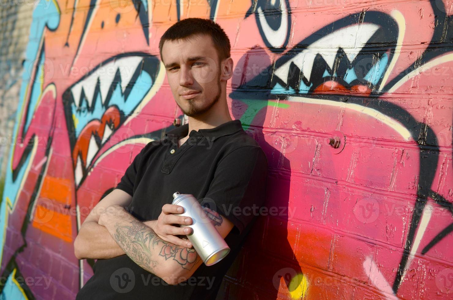 Young caucasian graffiti artist in black t-shirt with silver aerosol spray can near colorful graffiti in pink tones on brick wall. Street art and contemporary painting process photo