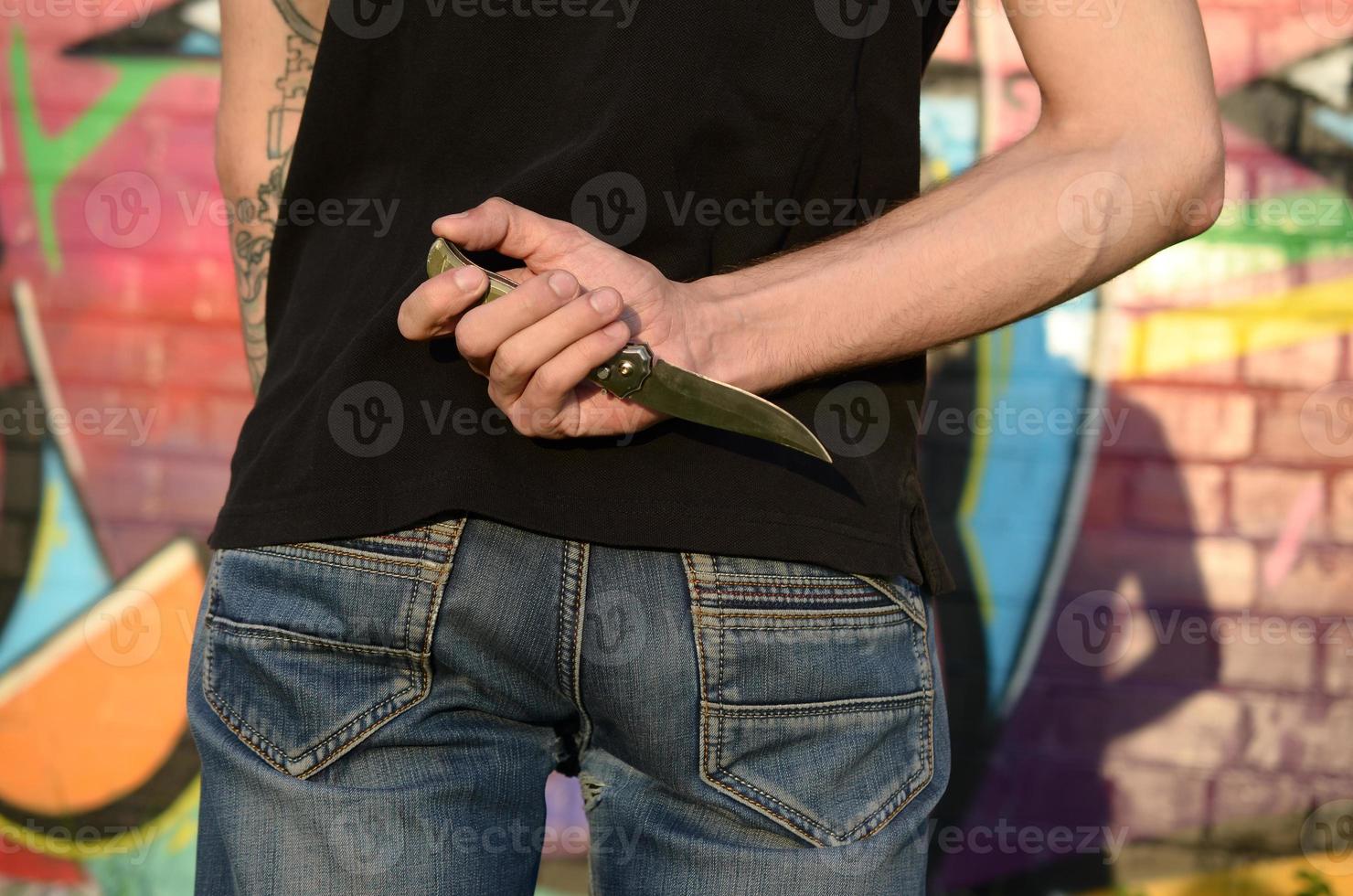 Back view of young caucasian man with knife in his hand against ghetto brick wall with graffiti paintings. Concept of criminal forces and aggression charge photo