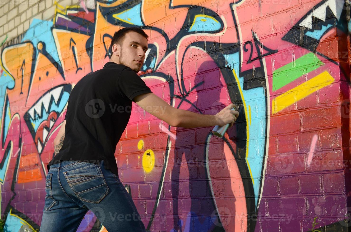 Young graffiti artist with backpack and gas mask on his neck paints colorful graffiti in pink tones on brick wall. Street art and contemporary painting process photo