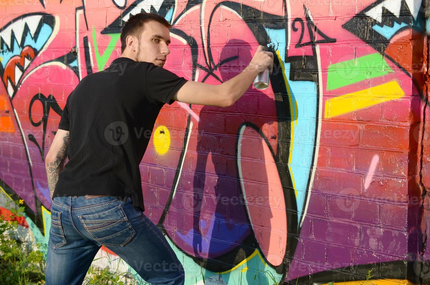 Young graffiti artist with backpack and gas mask on his neck paints colorful graffiti in pink tones on brick wall. Street art and contemporary painting process photo