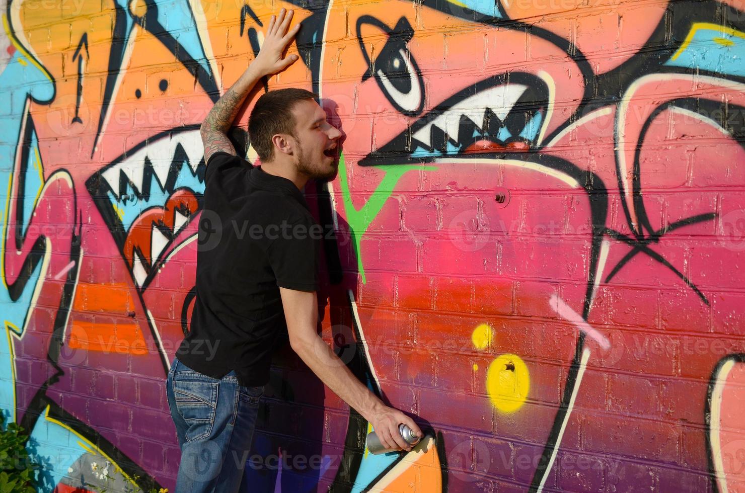 Young caucasian graffiti artist in black t-shirt with silver aerosol spray can near colorful graffiti in pink tones on brick wall. Street art and contemporary painting process photo
