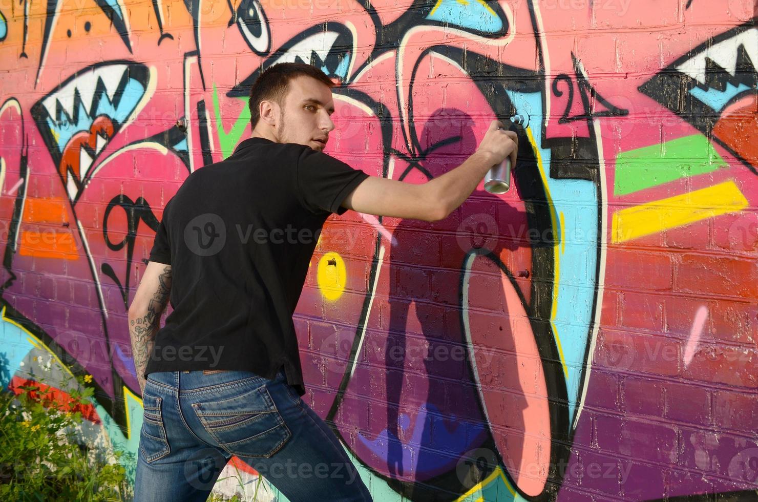 Young graffiti artist with backpack and gas mask on his neck paints colorful graffiti in pink tones on brick wall. Street art and contemporary painting process photo