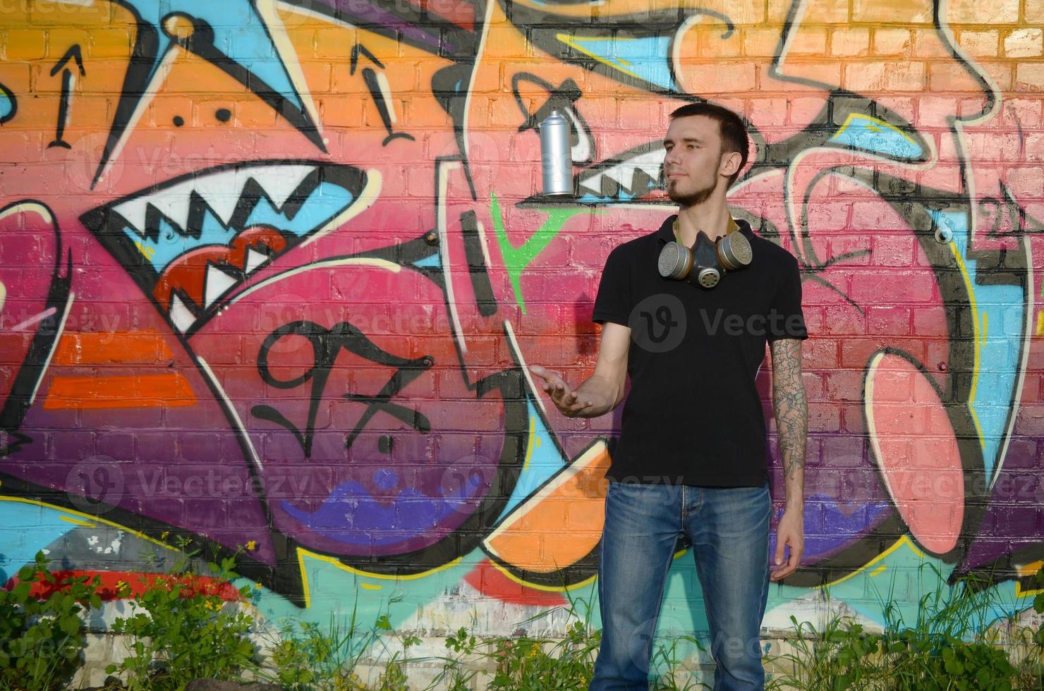 Young graffiti artist with gas mask on his neck throw his spray can against colorful pink graffiti on brick wall. Street art and contemporary painting process photo