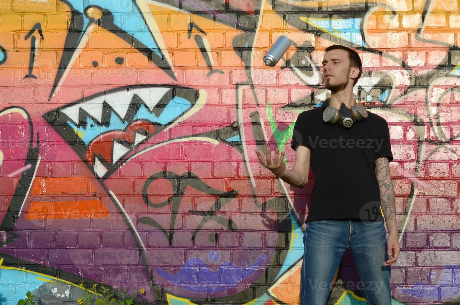 Young graffiti artist with gas mask on his neck throw his spray can against colorful pink graffiti on brick wall. Street art and contemporary painting process photo