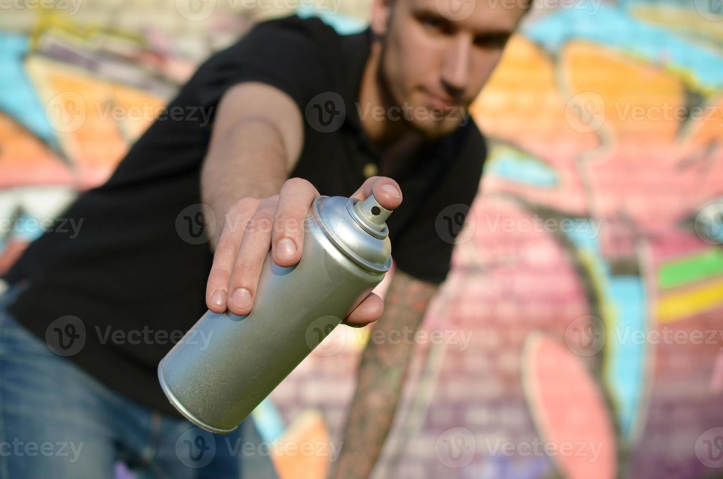 Young graffiti artist aims his spray can on background of colorful graffiti in pink tones on brick wall. Street art and contemporary painting process photo