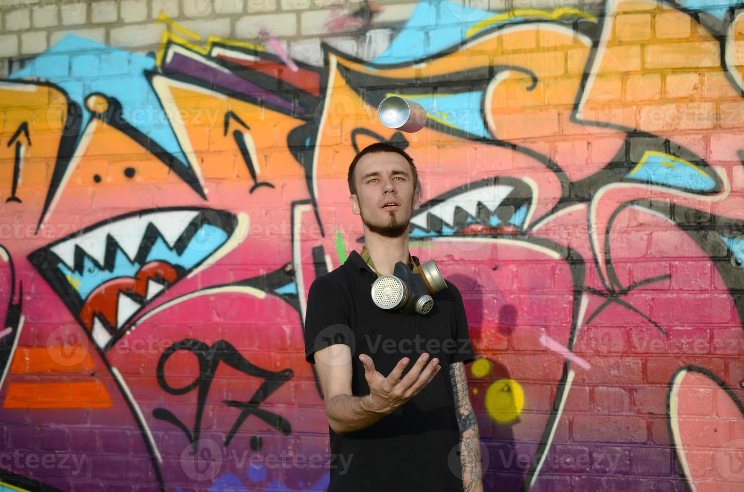 el joven artista de graffiti con máscara de gas en el cuello lanza su lata de aerosol contra el colorido graffiti rosa en la pared de ladrillo. proceso de arte callejero y pintura contemporánea foto