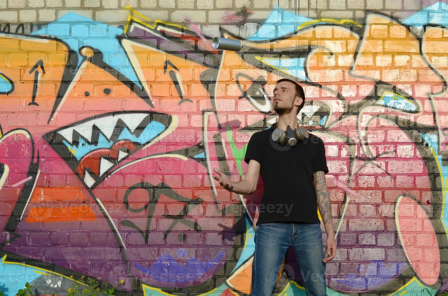 Young graffiti artist with gas mask on his neck throw his spray can against colorful pink graffiti on brick wall. Street art and contemporary painting process photo