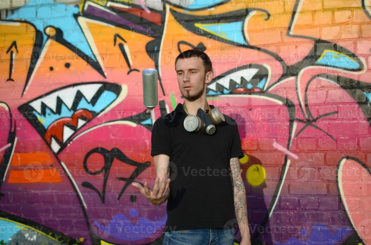 Young graffiti artist with gas mask on his neck throw his spray can against colorful pink graffiti on brick wall. Street art and contemporary painting process photo