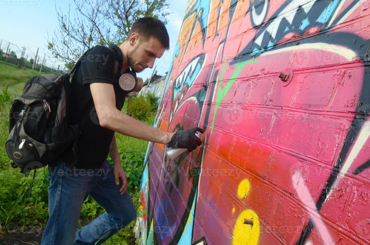 joven artista de graffiti con mochila y máscara de gas en el cuello pinta graffiti colorido en tonos rosas en la pared de ladrillo. proceso de arte callejero y pintura contemporánea foto