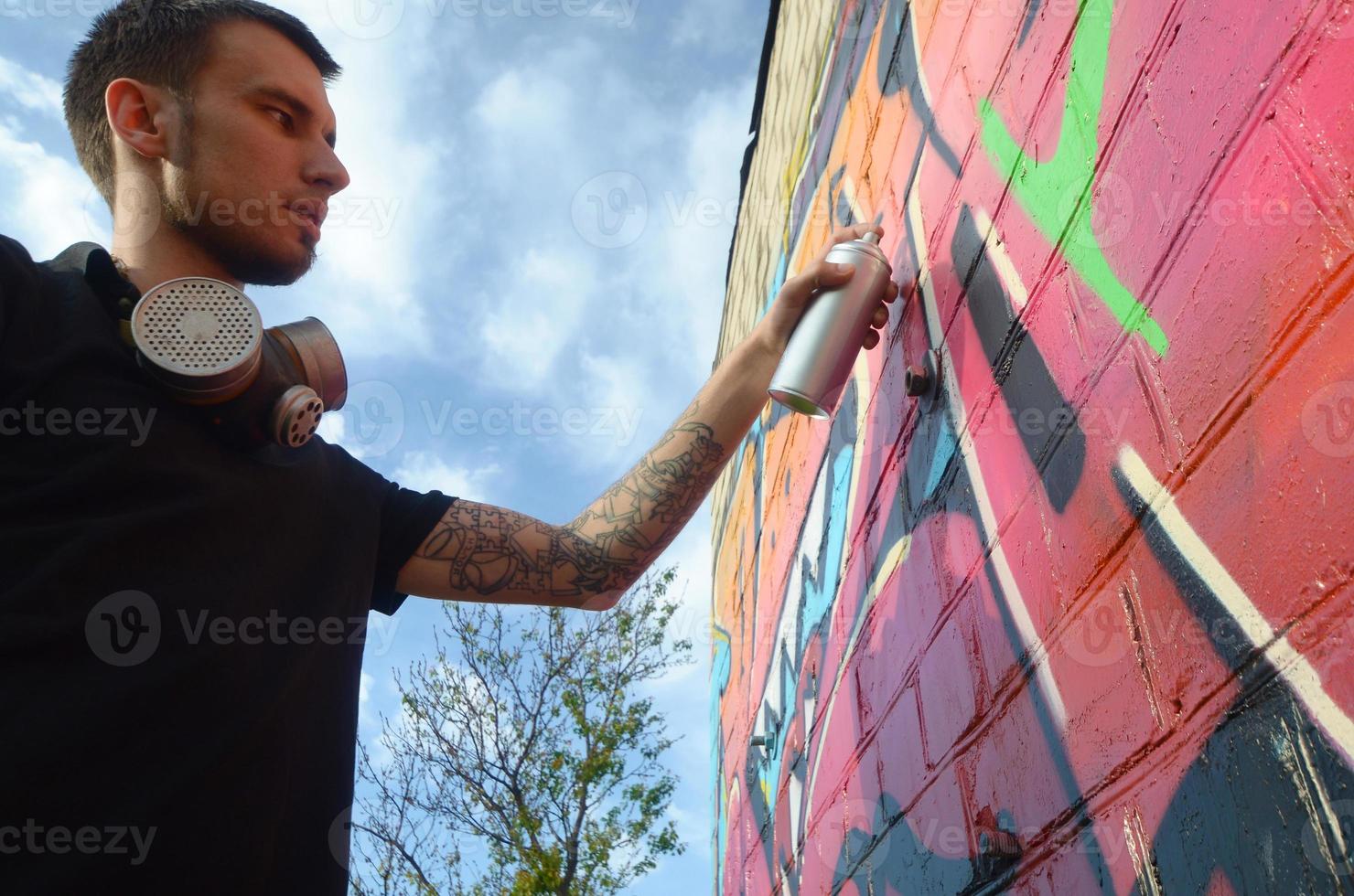 joven artista de graffiti con mochila y máscara de gas en el cuello pinta graffiti colorido en tonos rosas en la pared de ladrillo. proceso de arte callejero y pintura contemporánea foto